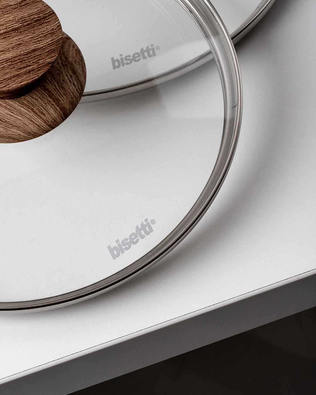 Two transparent glass saucepan lids with stainless-steel rims and wooden knobs, Bisetti logo visible, on a white countertop