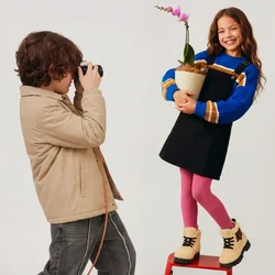 Ragazzo scatta una foto a una ragazza in piedi su un piccolo sgabello rosso che tiene in mano un'orchidea in vaso, entrambi sorridenti su uno sfondo semplice.