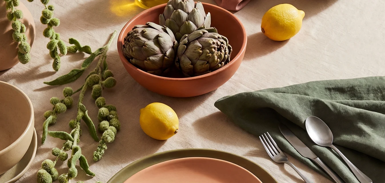 Tavolo rotondo con tovaglia beige, piatti in terracotta e oliva, carciofi in ciotola, fichi in ciotola, limoni, olio d'oliva, tovagliolo verde e sedia in legno.