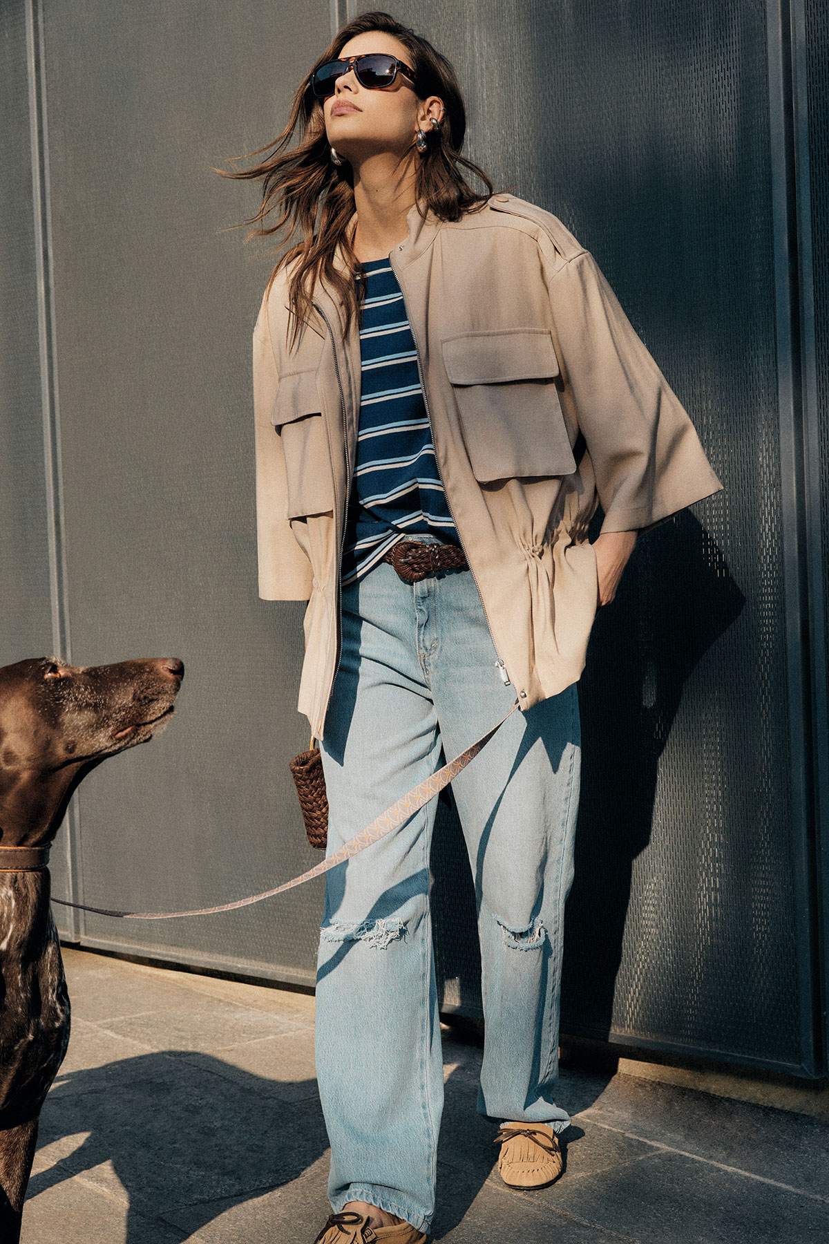 Woman in beige jacket, striped top and ripped jeans wearing sunglasses, walking a brown dog on a lead against an urban wall