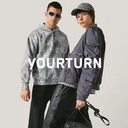 Due persone vestite alla moda posano davanti a uno sfondo neutro, indossando pantaloni scuri, top a strati e accessori come occhiali da sole e una borsa.