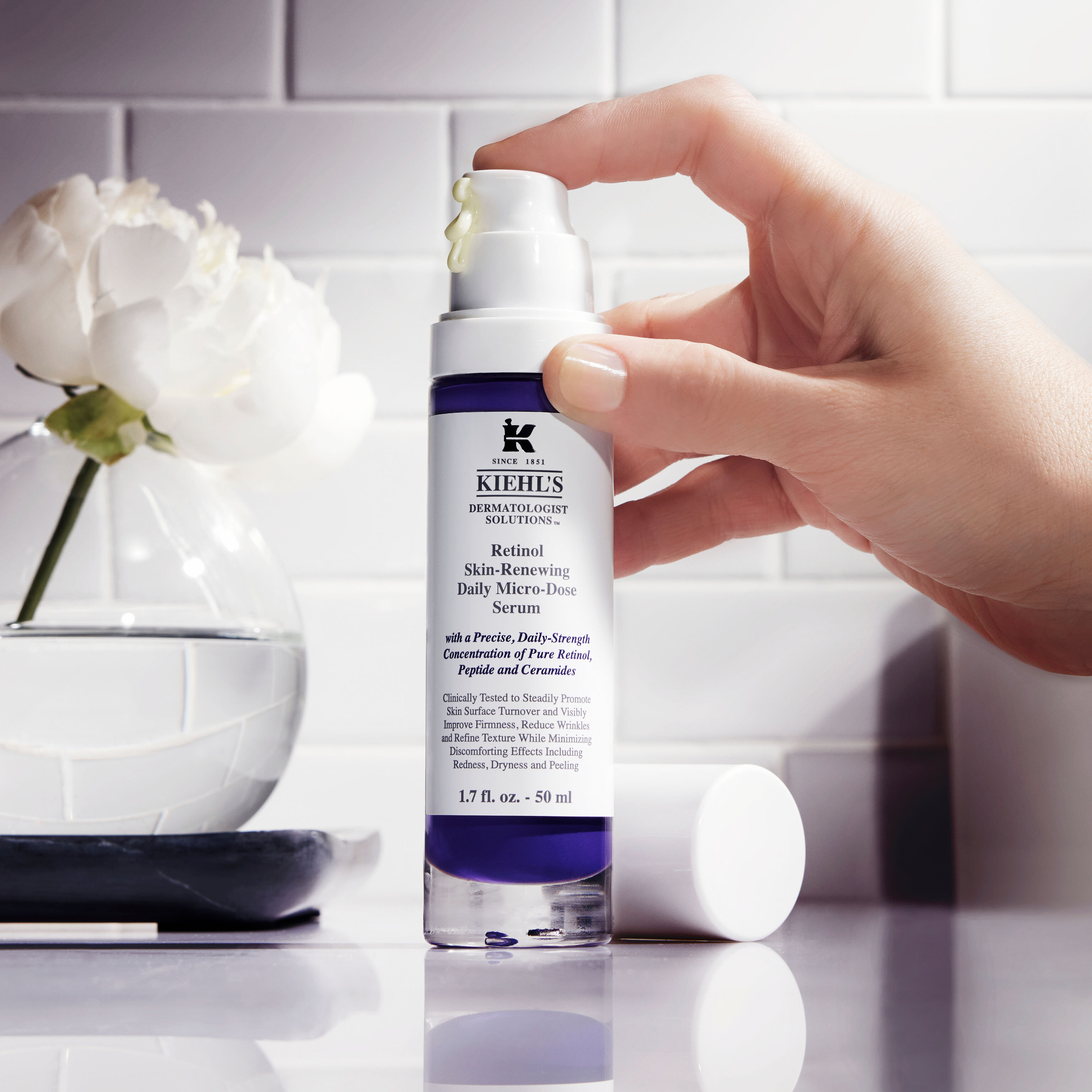 Una mano tiene il siero Retinol Daily Micro-Dose di Kiehl's davanti a un vaso di fiori bianchi
