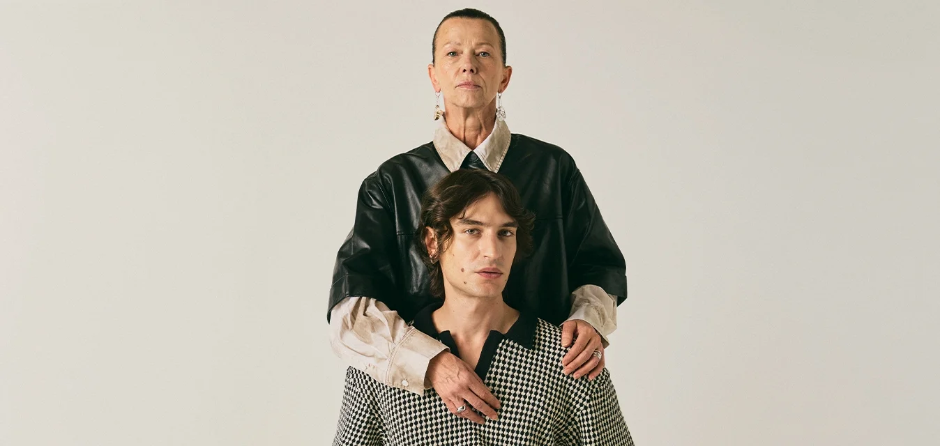 Due persone con espressioni serie posano; una sta in piedi dietro l'altra seduta, con le mani sulle spalle di quest'ultima, su uno sfondo semplice e chiaro.