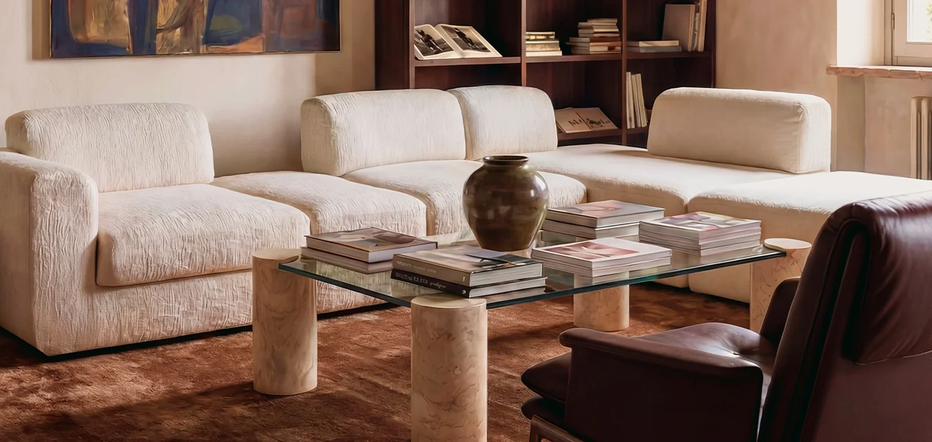 Soggiorno con divano angolare color crema, tavolino in vetro con libri e vaso, poltrona scura, libreria, opera d'arte e ulivo in vaso vicino alla finestra.