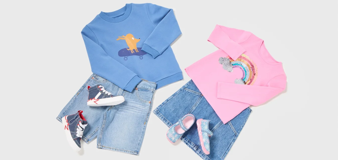 Maglione blu con cane sul monopattino, pantaloni corti in denim, sneakers blu navy; maglione rosa con arcobaleno, gonna in denim e scarpe rosa-blu, su sfondo bianco.