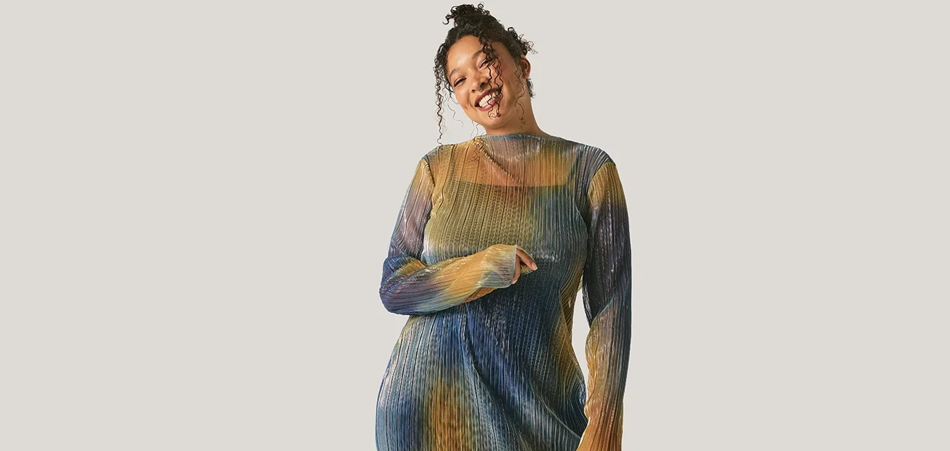 Giovane donna sorridente, con indosso un abito a rete multicolore e calze nere, in piedi contro uno sfondo chiaro uniforme.