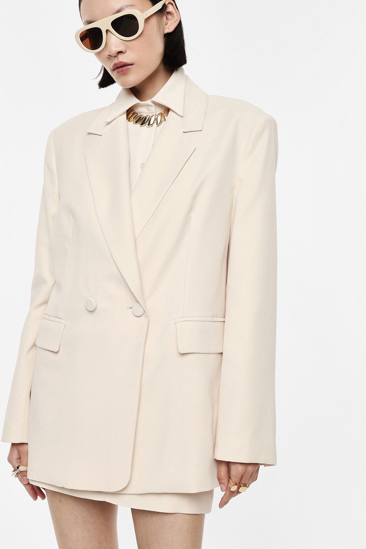 Woman modelling oversized cream blazer and mini skirt with white shirt, chunky gold necklace, cream sunglasses and rings