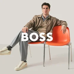 Uomo con blazer beige e pantaloni grigi seduto con le gambe incrociate su una delle due sedie arancioni collegate, su sfondo uniforme.