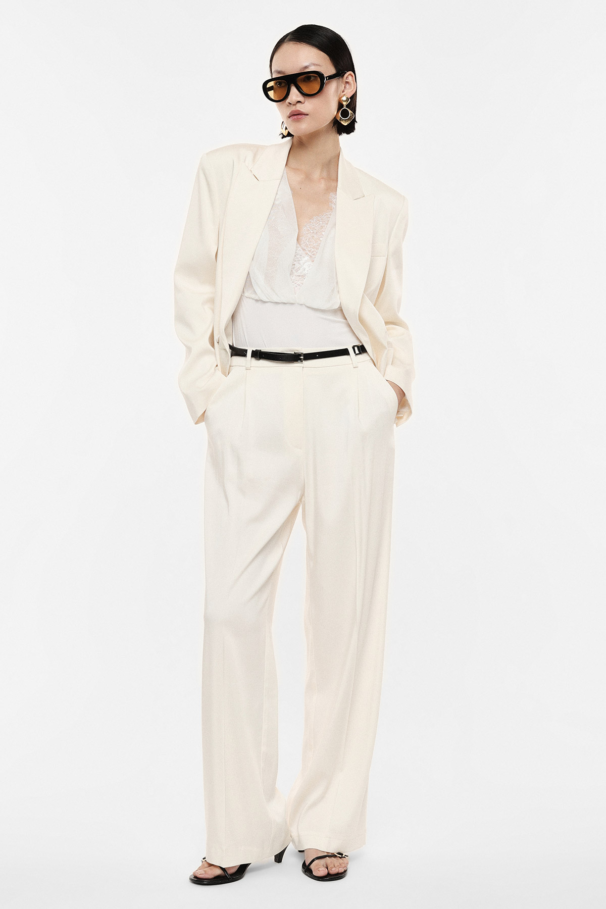 Woman in cream tailored suit and lace camisole with black belt, sunglasses and statement earrings on white background