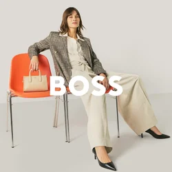 Donna in abito da lavoro seduta in modo rilassato su due sedie arancioni, con una borsa beige appoggiata su una delle sedie, su uno sfondo chiaro e uniforme.