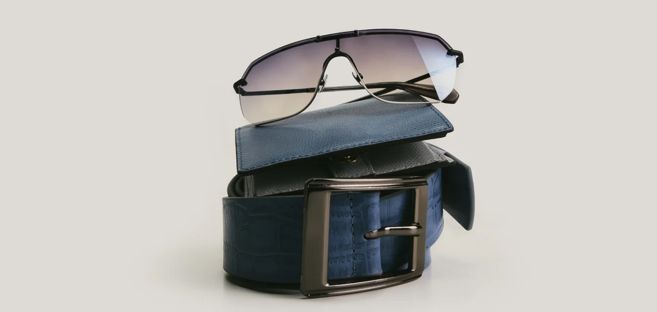 Cintura in pelle blu scuro piegata con fibbia in metallo sormontata da occhiali da sole neri con lenti sfumate su sfondo semplice.