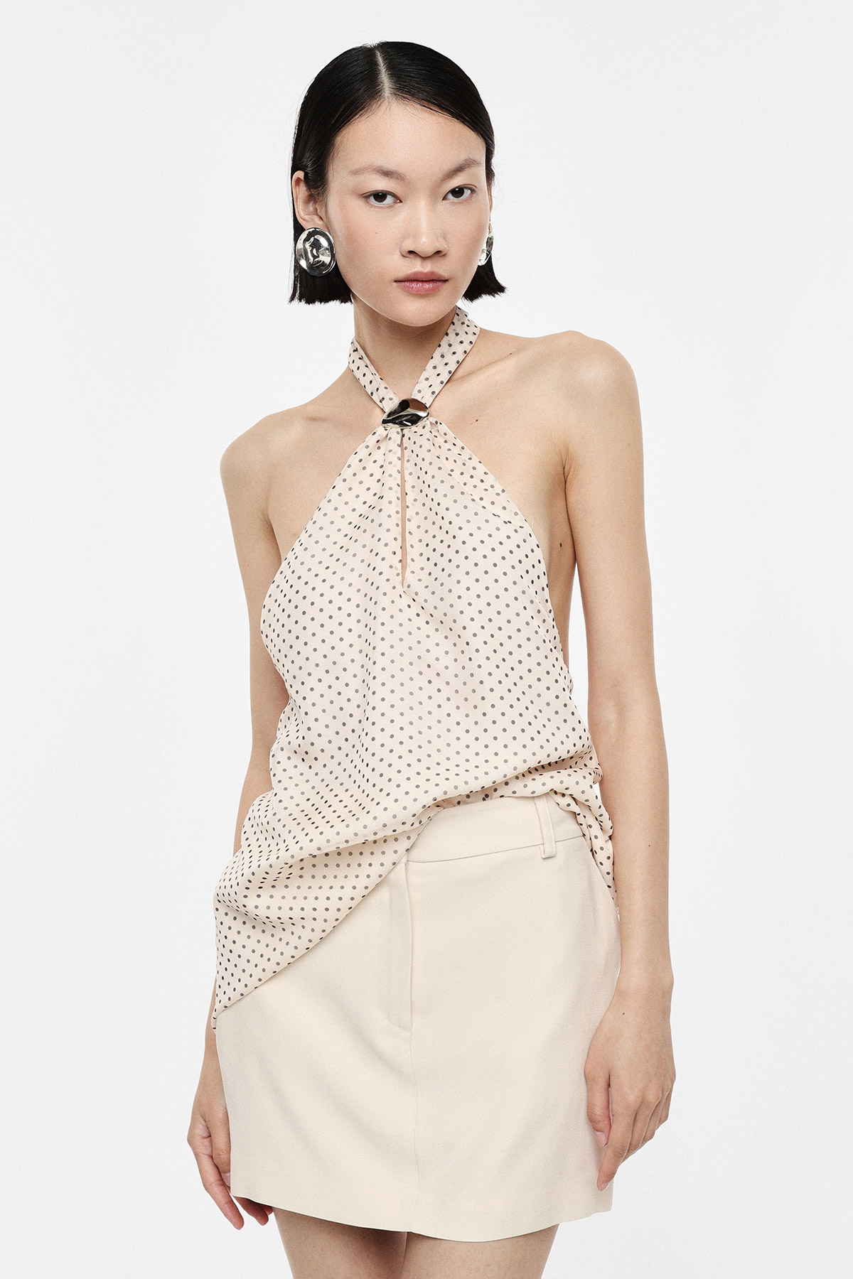 Model wearing cream polka-dot halter top with knot and cream mini skirt, silver statement earrings, neutral studio background