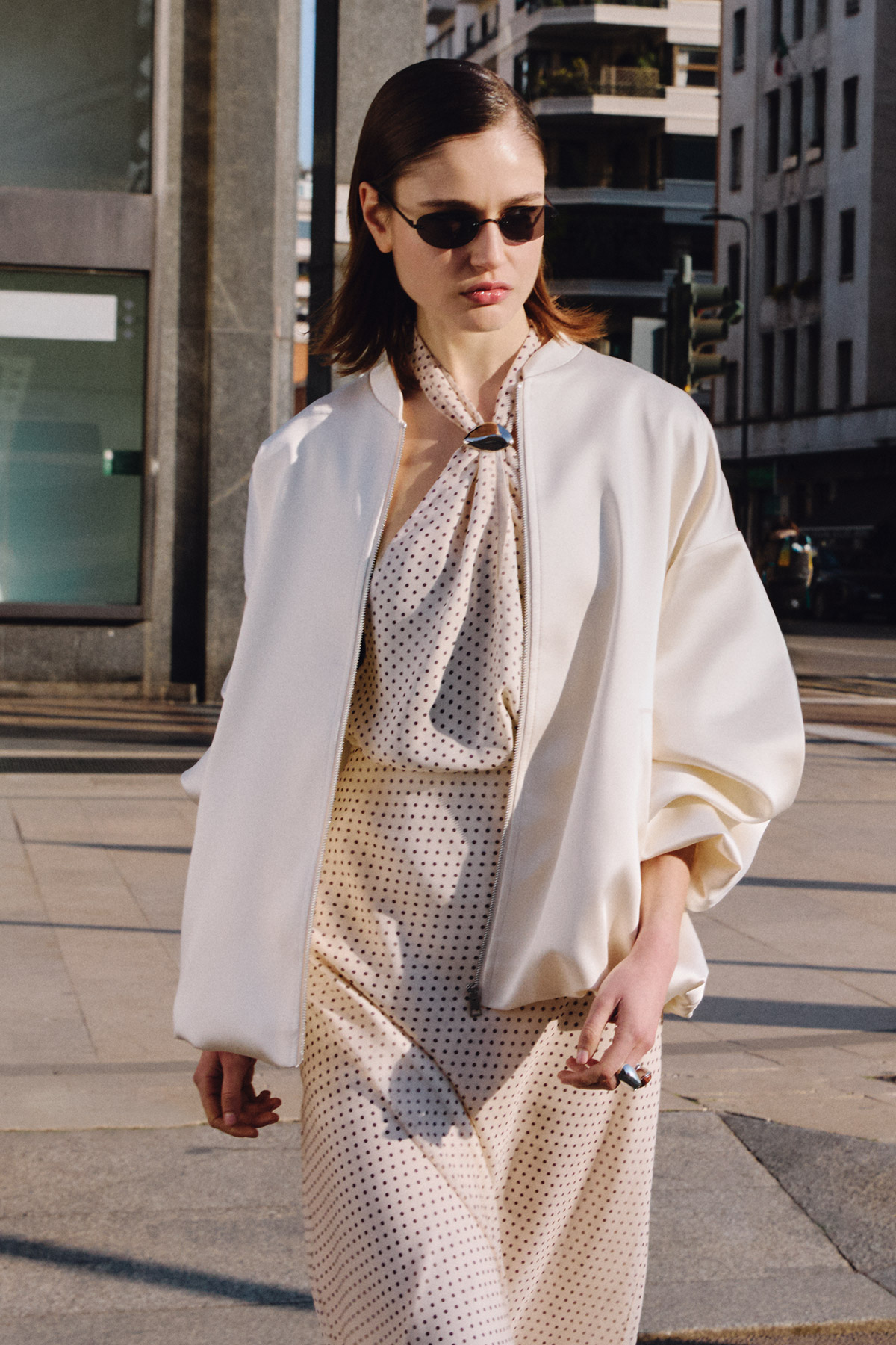 Woman in white bomber jacket, polka-dot silk dress and slim sunglasses walking a city street, chic street-style fashion