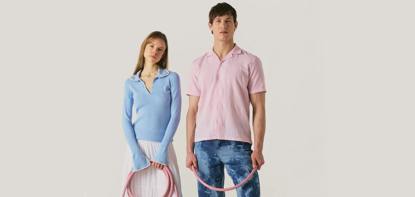 Donna con maglione blu e gonna bianca e uomo con camicia rosa e jeans blu tengono insieme un tubo rosa arrotolato, in piedi su sfondo chiaro.