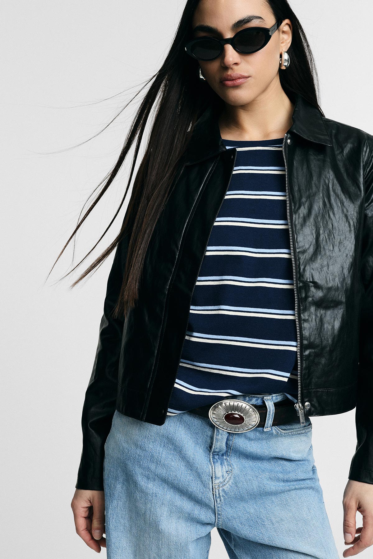 Woman wearing black leather jacket, navy striped top, oval sunglasses and light blue jeans with a silver concho belt buckle
