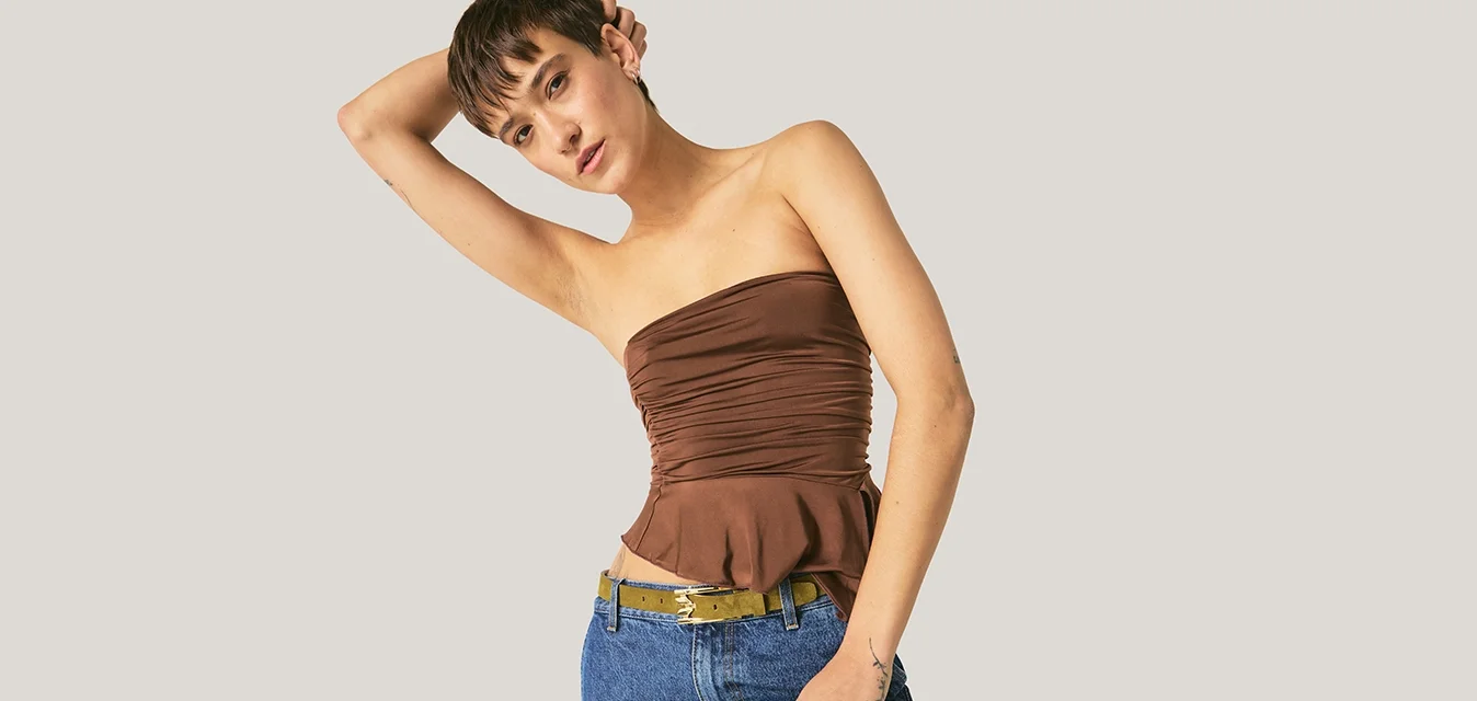 Donna con top marrone senza spalline e jeans blu si inclina da un lato, appoggiando un braccio sulla testa contro uno sfondo uniforme.