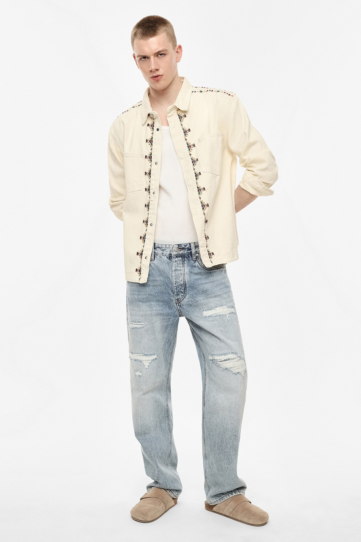 Male model wearing embroidered cream overshirt over white vest, distressed light-blue jeans and tan slippers, standing on white background