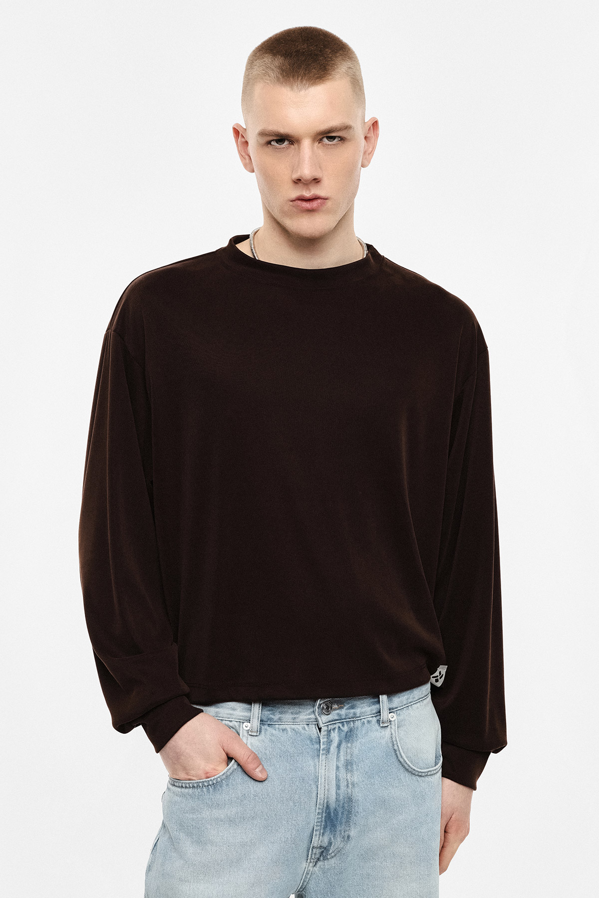 Young man in dark brown long-sleeved shirt and light blue jeans standing against white background with hand in pocket