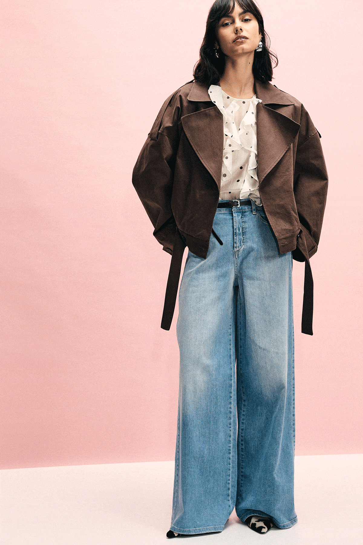 Three-shot of model wearing wide-leg light-blue jeans, cream polka-dot blouse and brown jacket on pink background