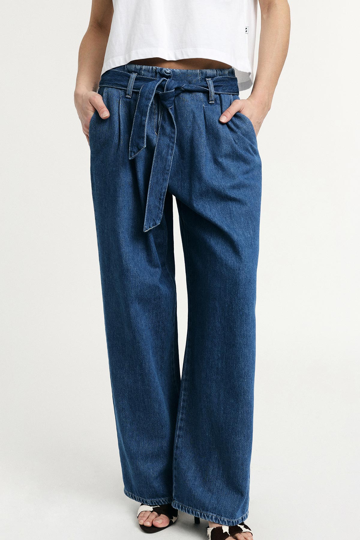 Woman wearing high-waisted wide-leg blue denim trousers with tie belt and pockets, styled with a white cropped top