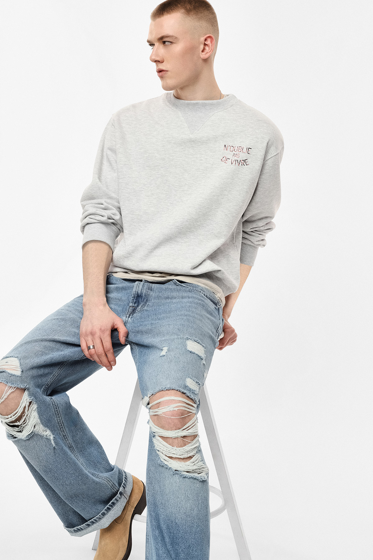 Young man sitting on stool in grey sweatshirt and ripped blue jeans with tan boots, looking left — casual men's fashion outfit