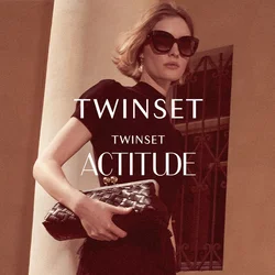 Donna con vestito nero e occhiali da sole tiene una pochette mentre cammina su gradini di pietra accanto a una colonna e una statua classica.