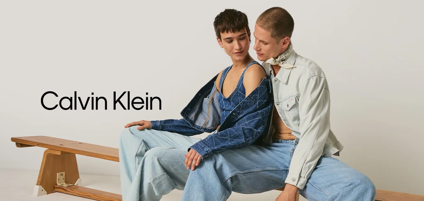 Due giovani adulti scalzi con indosso abbigliamento in denim sono seduti vicini su una panchina di legno davanti a uno sfondo semplice.