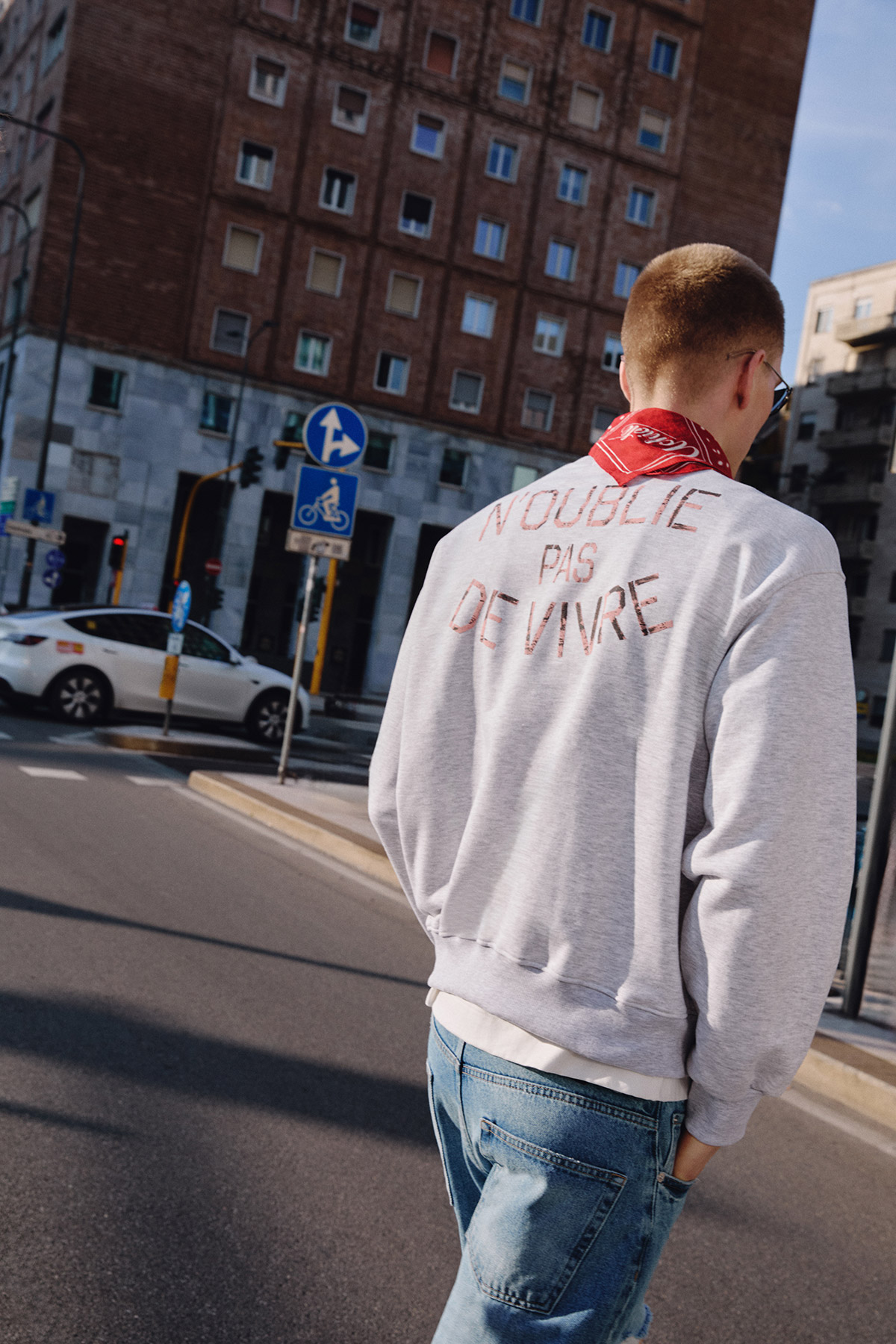 Man walking on urban street wearing grey sweatshirt reading 'N'oublie pas de vivre' and red bandana, buildings and traffic
