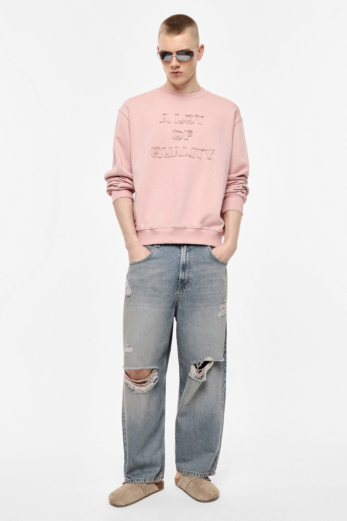 Young man in pink sweatshirt reading 'A LOT OF QUALITY', ripped wide-leg jeans, sunglasses and beige slippers