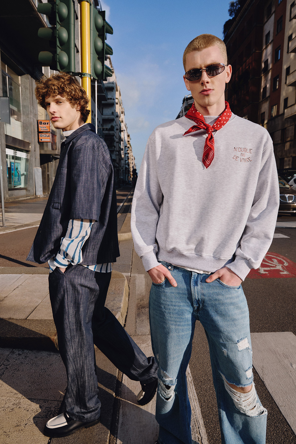 Two young male fashion models on an urban street: one in a grey sweatshirt with a red bandana and ripped jeans, the other in a dark suit.