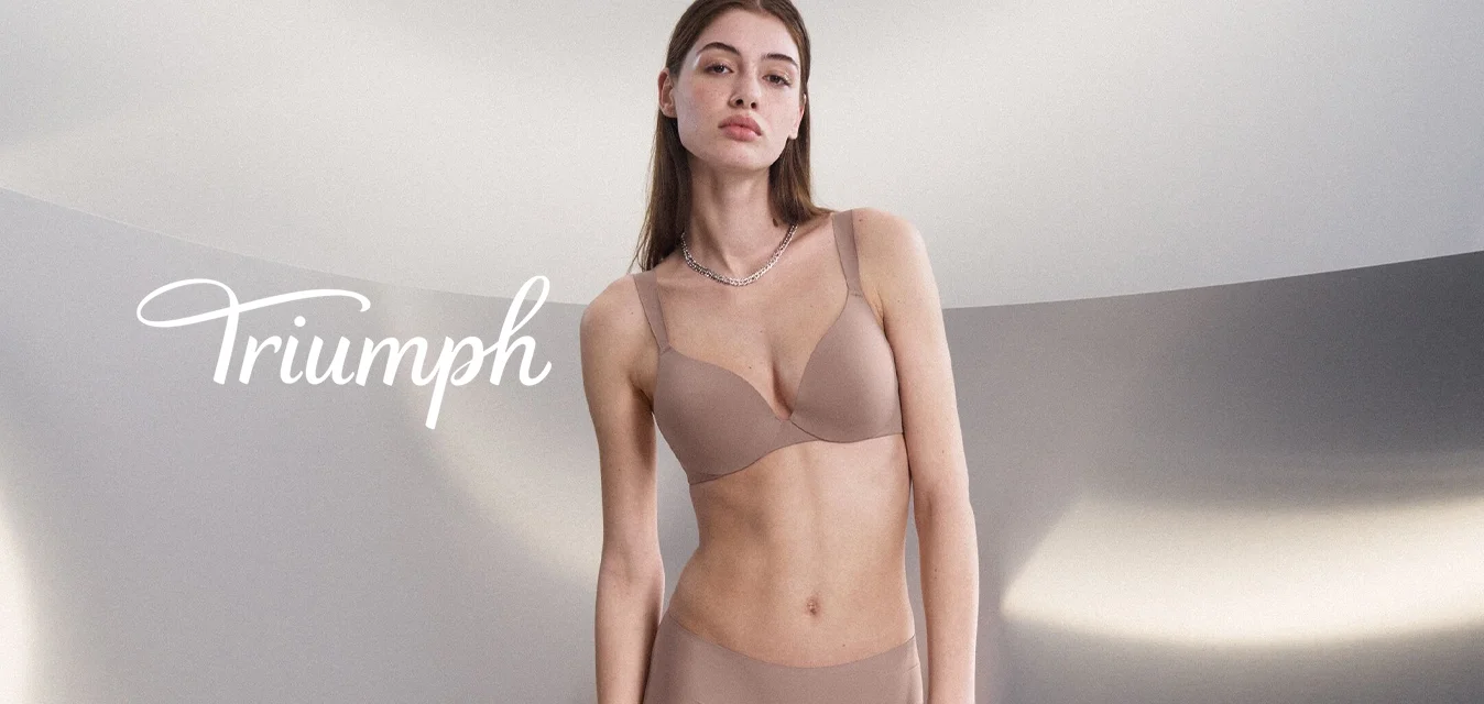 Giovane donna con reggiseno e slip beige, in piedi e che tiene un capo beige contro uno sfondo grigio chiaro minimalista e curvo.