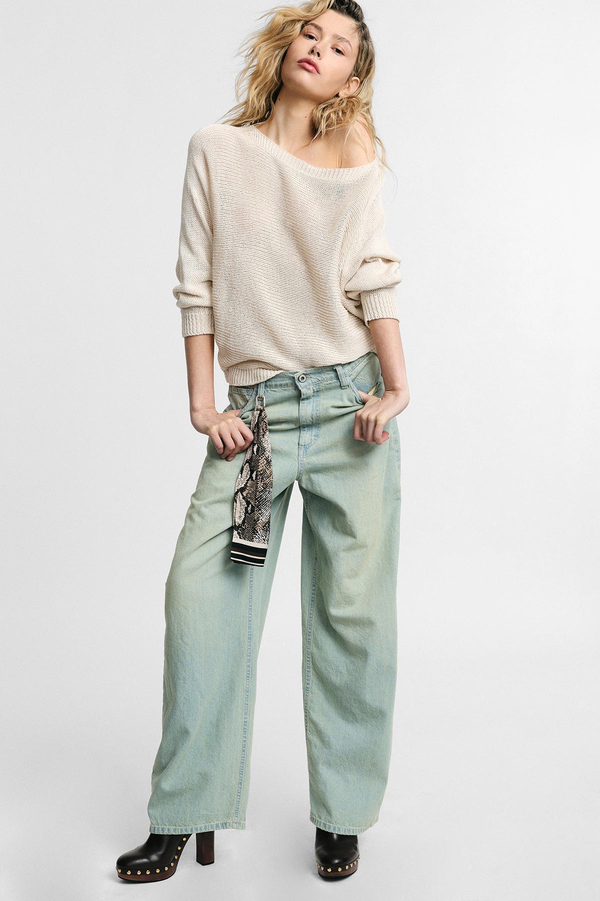 Model wearing cream off-shoulder knit sweater, light-wash wide-leg jeans with hanging patterned scarf and black studded platform boots