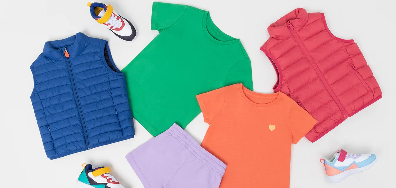 Un set di abbigliamento colorato per bambini include un gilet blu gonfiabile, t-shirt verdi e arancioni, pantaloni corti viola e sneakers colorate con un design.
