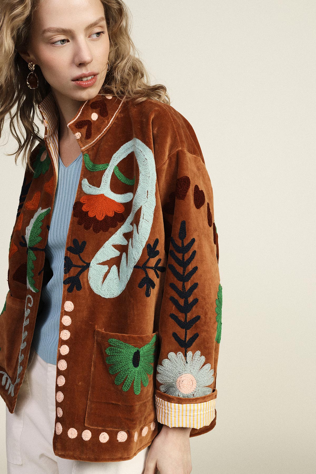 Woman wearing brown suede embroidered jacket with colourful floral and leaf motifs, blue knit top, looking left