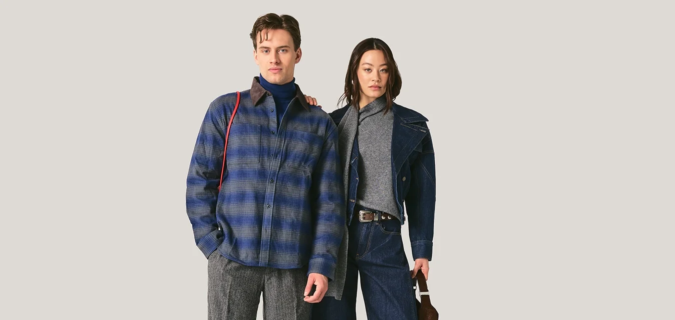 Uomo con giacca a quadri blu e pantaloni grigi accanto a donna con giacca scura, dolcevita grigio, pantaloni larghi blu navy e tacchi neri che tiene una piccola borsa.