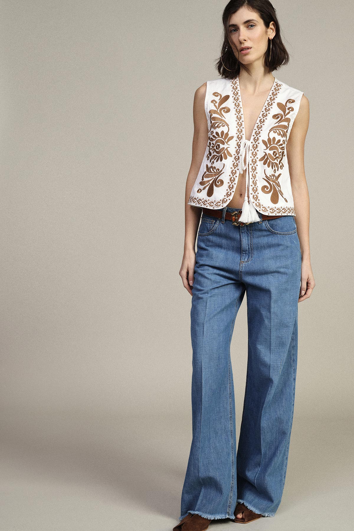 Model wearing a white embroidered sleeveless vest with tassel tie and high-waist wide-leg blue jeans against a neutral background