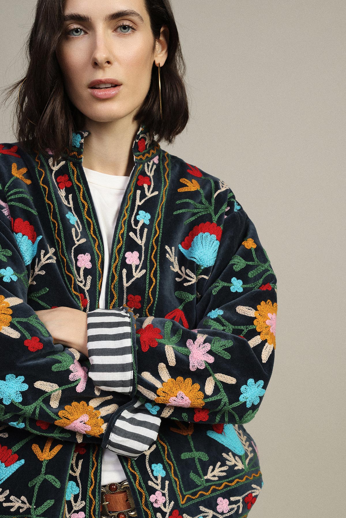 Woman wearing embroidered floral velvet jacket with striped cuff, gold hoop earring, arms folded against neutral background