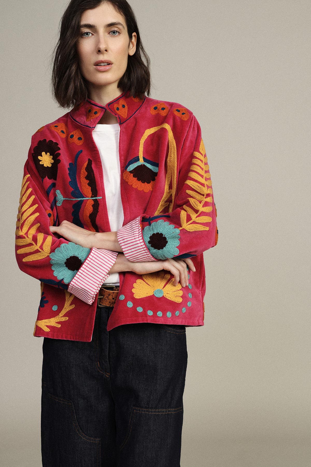Woman wearing embroidered red floral jacket over white tee, dark jeans, arms crossed against neutral background