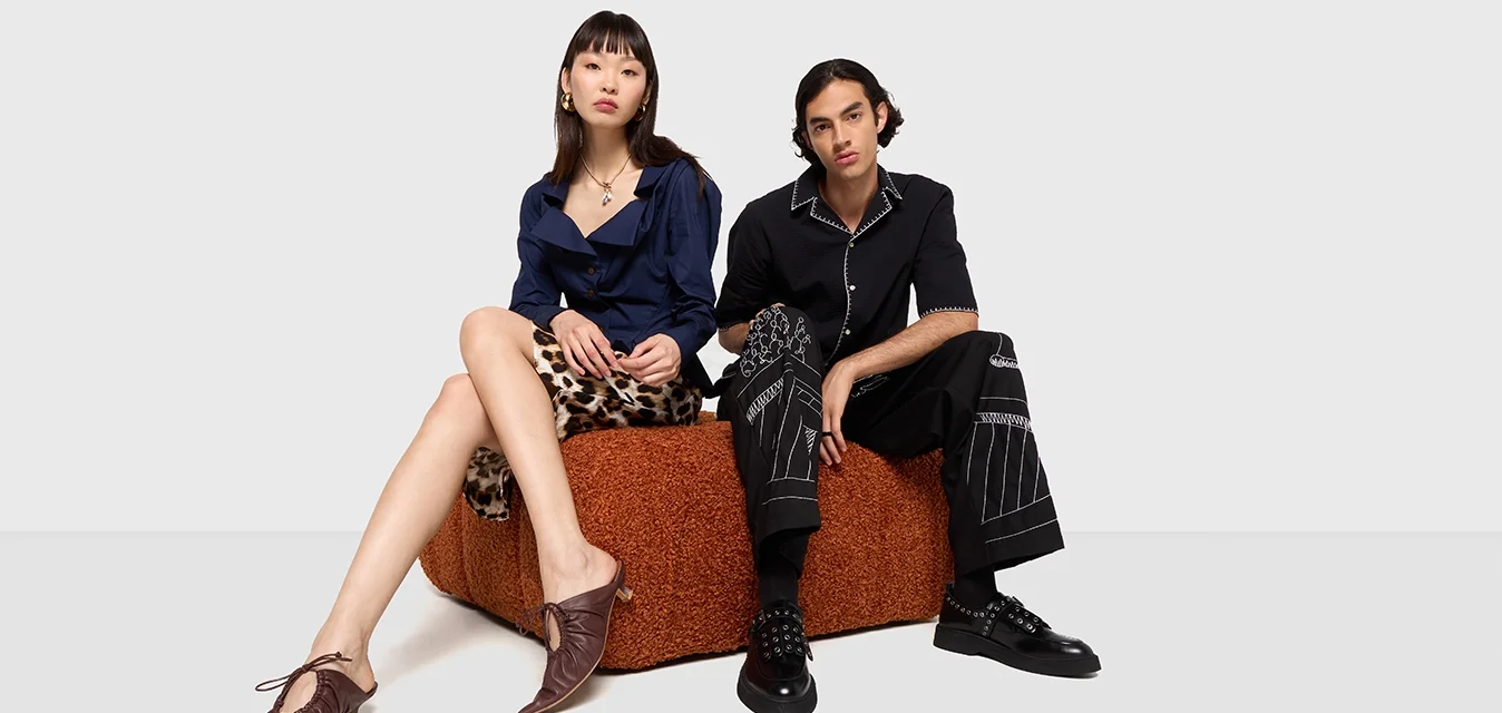 Due persone sono sedute su un pezzo di arredamento marrone strutturato. Uno indossa una camicia blu e pantaloni a fantasia; l'altro, una maglietta nera e pantaloni a righe.