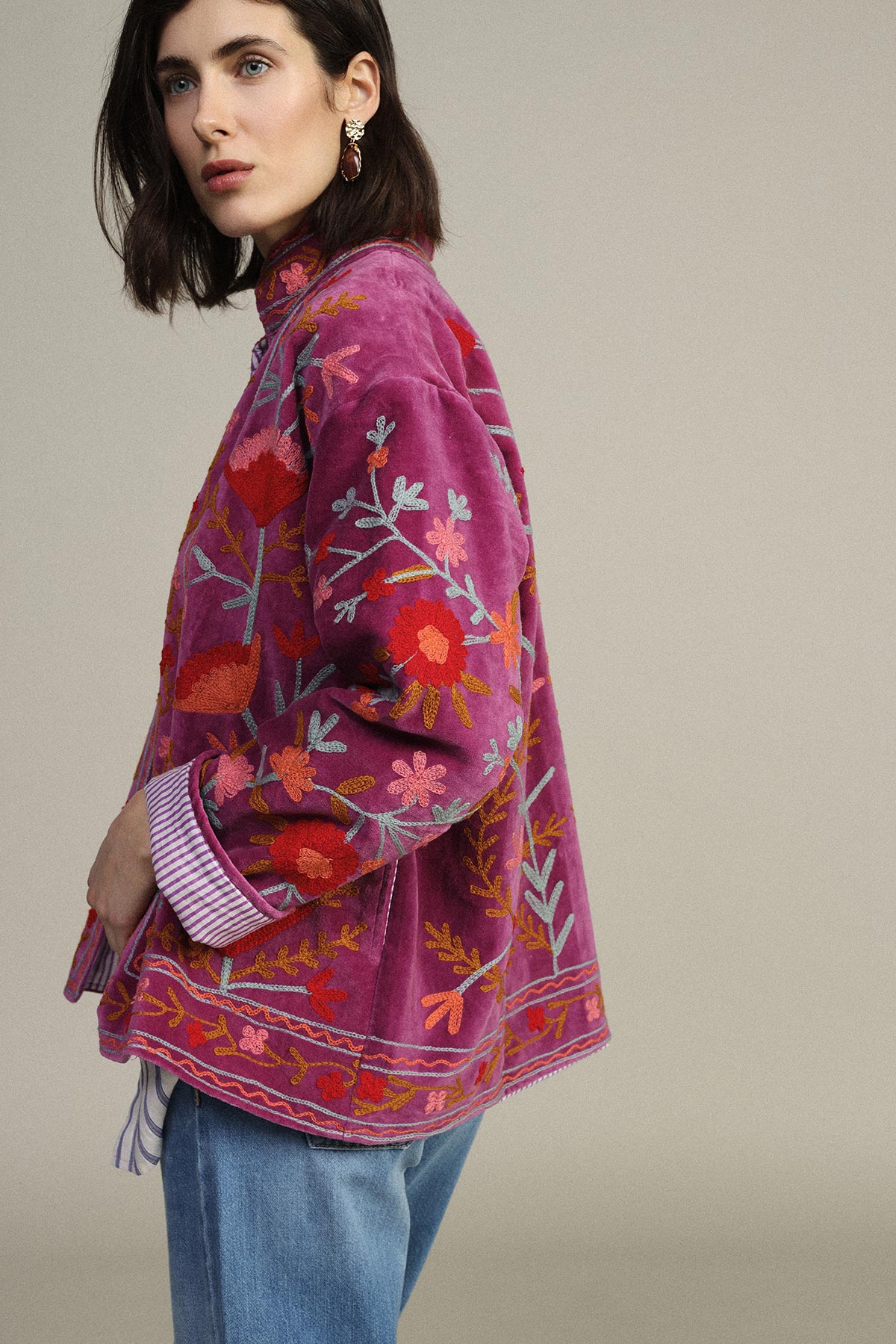 Woman wearing a magenta embroidered floral jacket and blue jeans, side profile on neutral background, gold drop earring