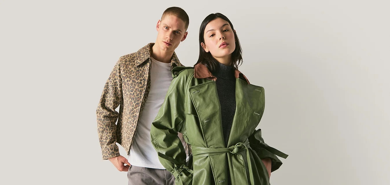 Donna con trench verde con cintura e top nero accanto a uomo con giacca leopardata, t-shirt bianca e pantaloni grigi su sfondo neutro.