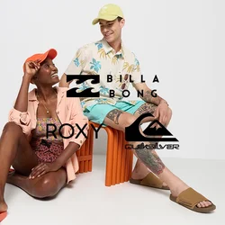 Due persone sono sedute in un ambiente minimalista. Una indossa un cappello arancione, una maglietta rosa chiaro e un costume da bagno a fantasia; l'altra ha un cappello verde chiaro, una camicia floreale e pantaloni corti. Entrambi indossano sandali.