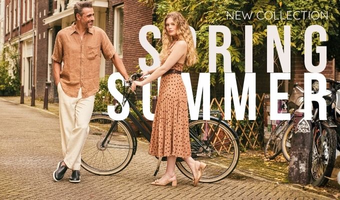 Man and woman in spring-summer outfits standing with a bicycle on a sunlit city street, outdoor fashion collection