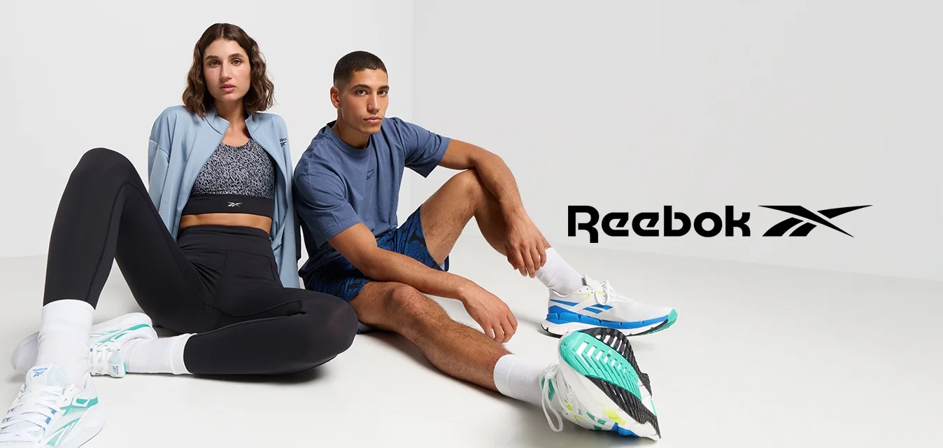 Due persone sono sedute su un pavimento bianco. Una indossa una giacca azzurro chiaro, leggings neri e sneakers bianche. L'altra indossa una maglietta blu e pantaloni corti.