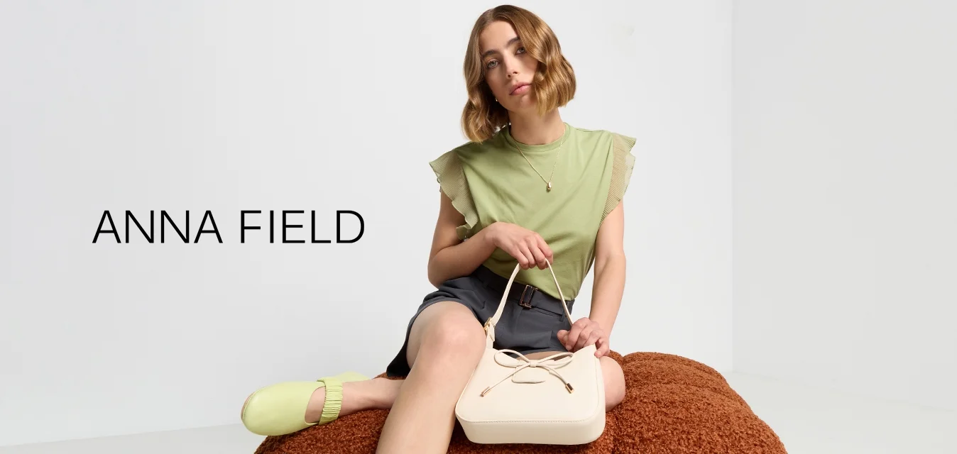 Borsa beige con dettaglio a nodo, abbinata a una maglietta verde e a pantaloni grigi. Il modello è seduto su un cuscino marrone texturizzato. Illuminazione soffusa.