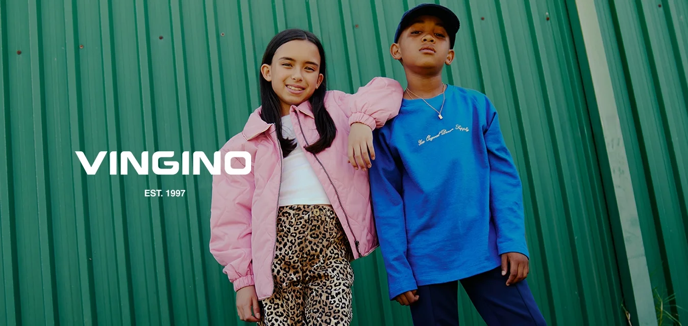 Due bambini sono in piedi sull'erba davanti a un muro di metallo verde; uno indossa una giacca rosa e pantaloni leopardati, l'altro una felpa blu e pantaloni scuri.