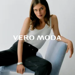 Donna con lunghi capelli castani seduta su una sedia moderna azzurro chiaro, indossa una canottiera bianca e jeans neri a gamba larga su sfondo bianco semplice.