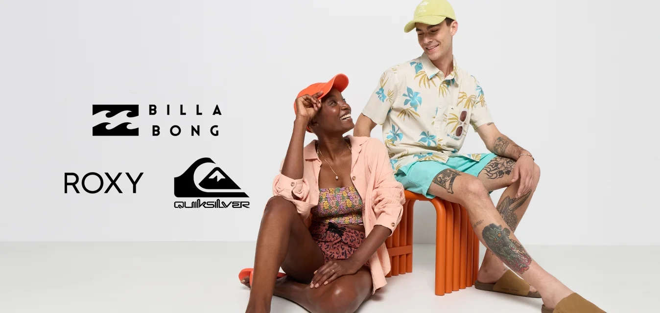 Due persone sono sedute in un ambiente minimalista. Una indossa un cappello arancione, una maglietta rosa chiaro e un costume da bagno a fantasia; l'altra ha un cappello verde chiaro, una camicia floreale e pantaloni corti. Entrambi indossano sandali.