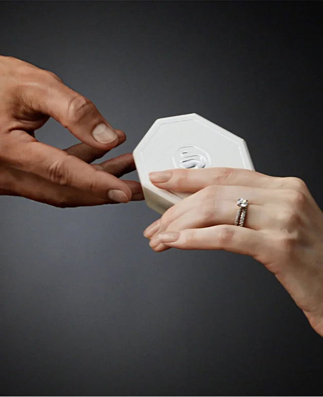 Mano che indossa un anello realizzato con Swarovski Created Diamonds e porge una confezione regalo Swarovski bianca ad un’altra mano