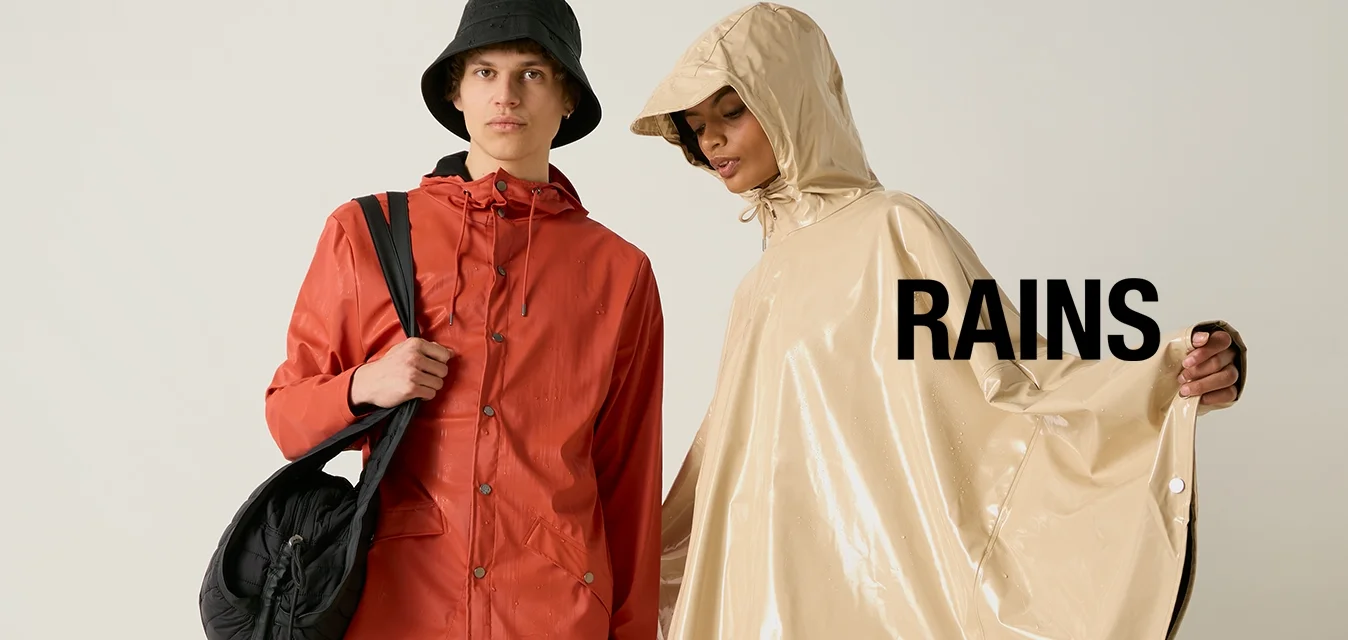 Due persone con abbigliamento impermeabile: una con giacca rossa e cappello a tesa larga nero con una borsa, l'altra con poncho beige con cappuccio e pantaloncini neri.